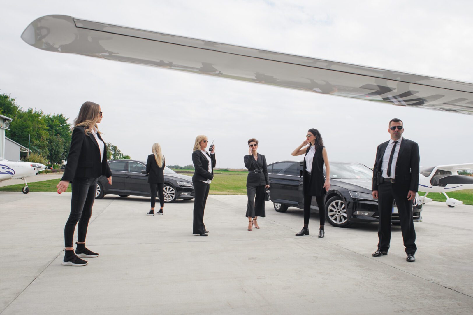 A group of professionally dressed people standing apart in an outdoor setting with cars.