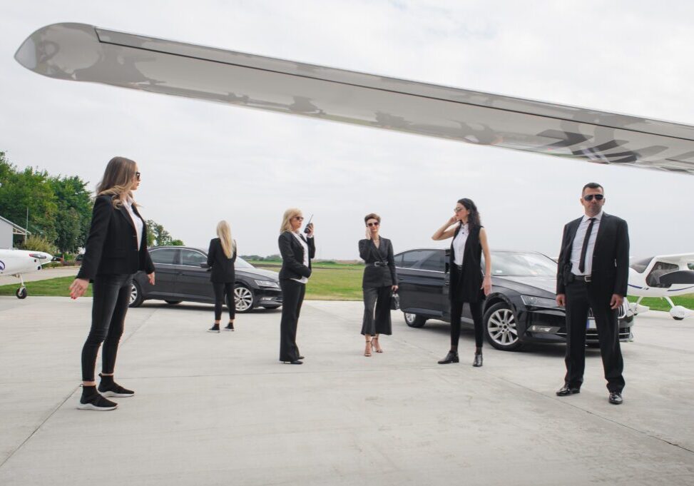 A group of professionally dressed people standing apart in an outdoor setting with cars.